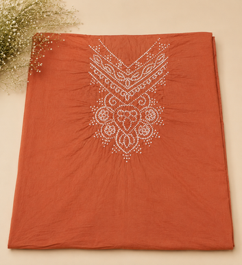 A rust-orange cotton fabric, likely a kurta or sari, featuring a white embroidered mandala pattern on the front. The fabric is neatly folded and presented on a light beige background with dried baby's breath flowers in the top-left corner.