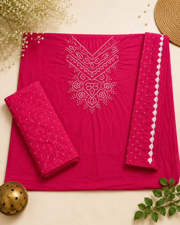 A bright pink salwar suit set laid flat on a light beige surface, featuring a kurta with white embroidered neckline details, matching folded pants, and a coordinating dupatta with white geometric border embroidery. Decorative flowers, a woven mat, and a small brass ornament frame the scene.