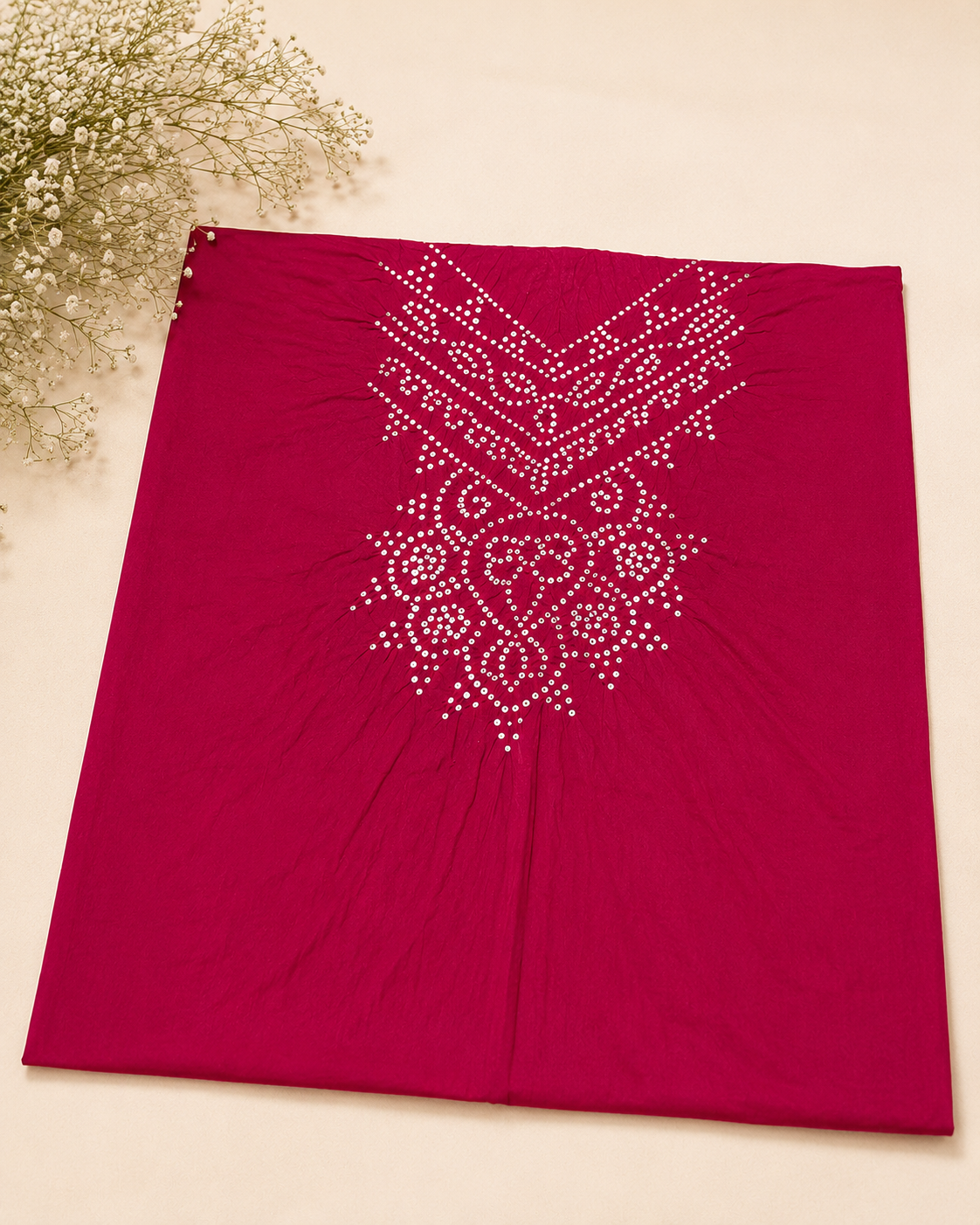 A folded deep magenta pink fabric garment, likely a kurta or dress, featuring a white embroidered geometric and floral pattern on the chest. The fabric is laid flat on a light beige background with a decorative sprig of baby's breath in the top-left corner.