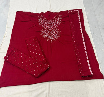 A folded deep red salwar suit set laid flat on a white fabric background, featuring white embroidered patterns on the chest, white polka dots on the fabric, and a white scalloped border on the dupatta.