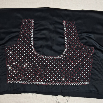 Black fabric blouse piece with mirror work embellishments and white zigzag border stitching, laid flat on a light beige surface.