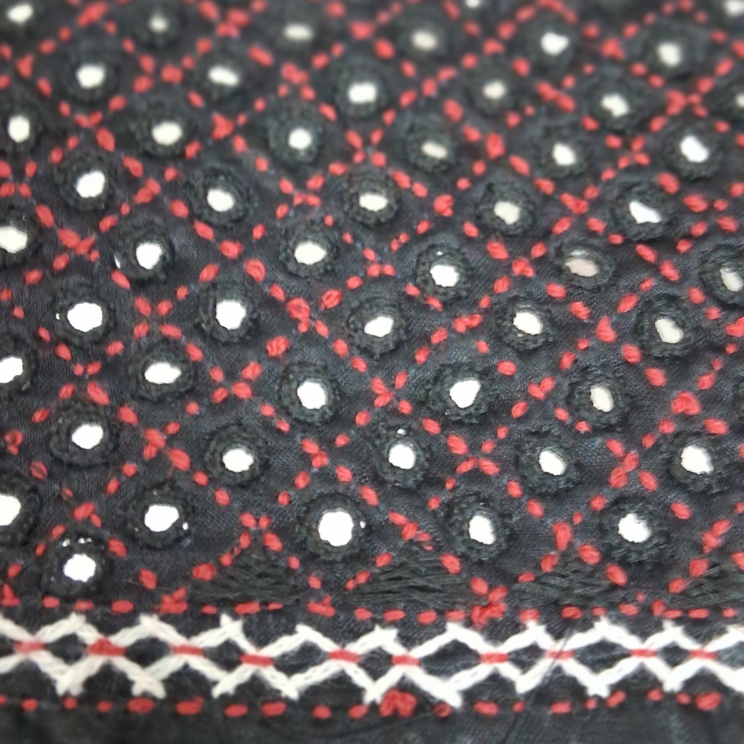 Close-up of a black fabric with red dotted geometric patterns and white circular embroidered motifs, plus a white zigzag border along the bottom.