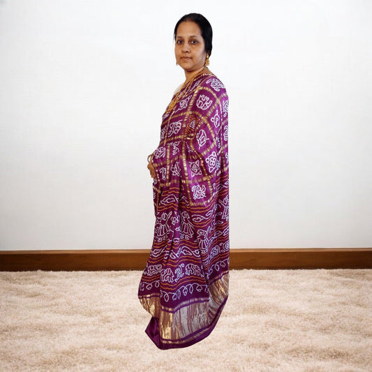 Woman wearing a purple bandhani saree saree standing against a plain white wall.
