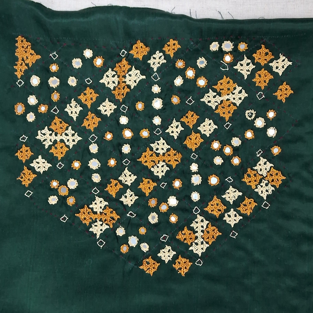 A close-up of a dark green textile featuring a large embroidered geometric pattern in orange and cream, with small scattered mirror work reflections.