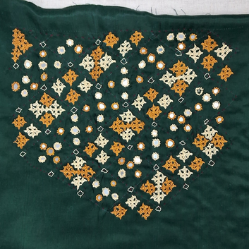 A close-up of a dark green textile featuring a large embroidered geometric pattern in orange and cream, with small scattered mirror work reflections.