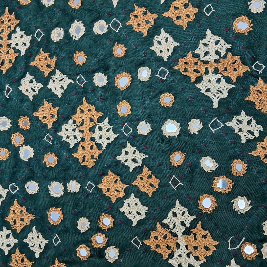 A close-up of a dark teal fabric featuring intricate hand-embroidered geometric and floral patterns in beige, mustard yellow, and white, accented with small iridescent mirror-like studs.