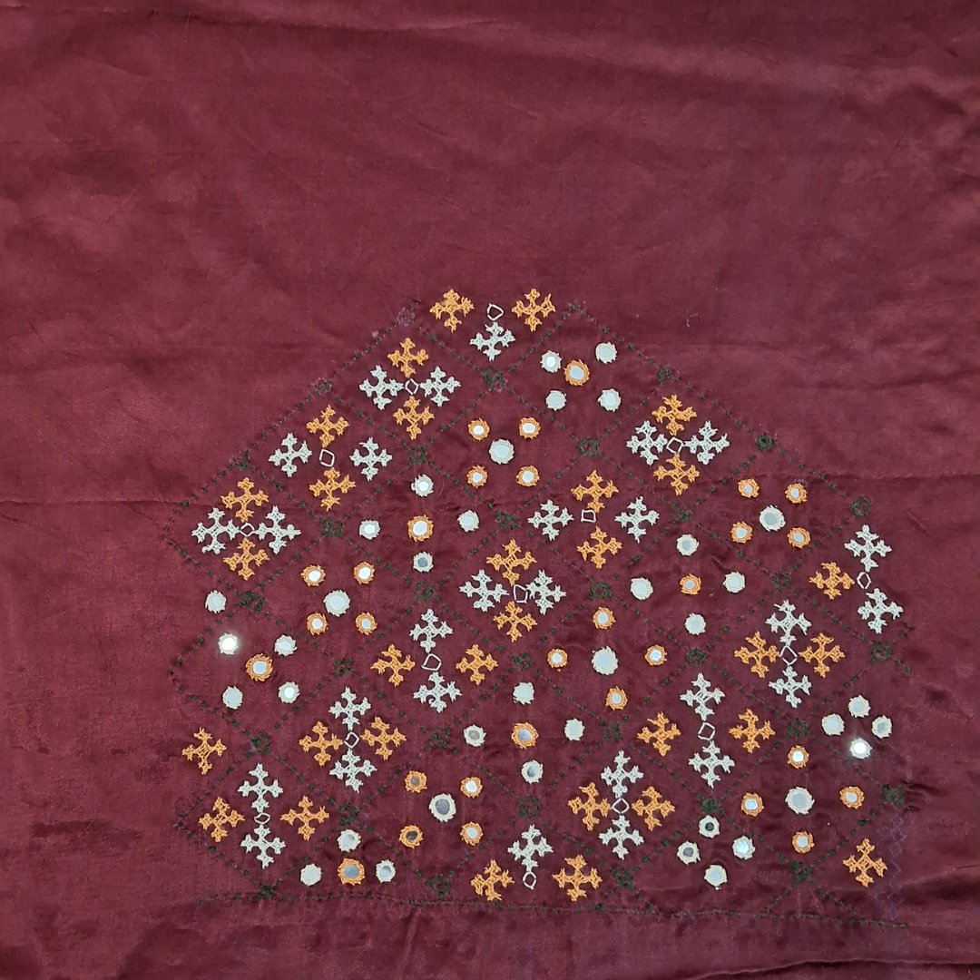 A close-up of a burgundy fabric surface with a decorative embroidered or printed geometric floral pattern in orange, white, and green arranged in a diamond-like cluster near the center.