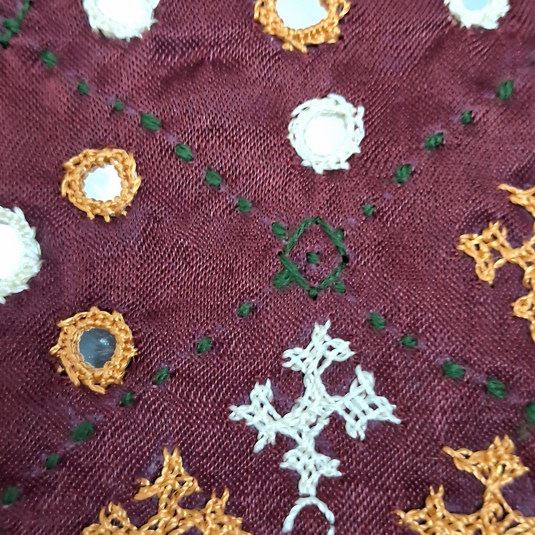 Close-up of a burgundy embroidered fabric with white and gold floral motifs, green dotted accents, and a central green diamond embroidery.