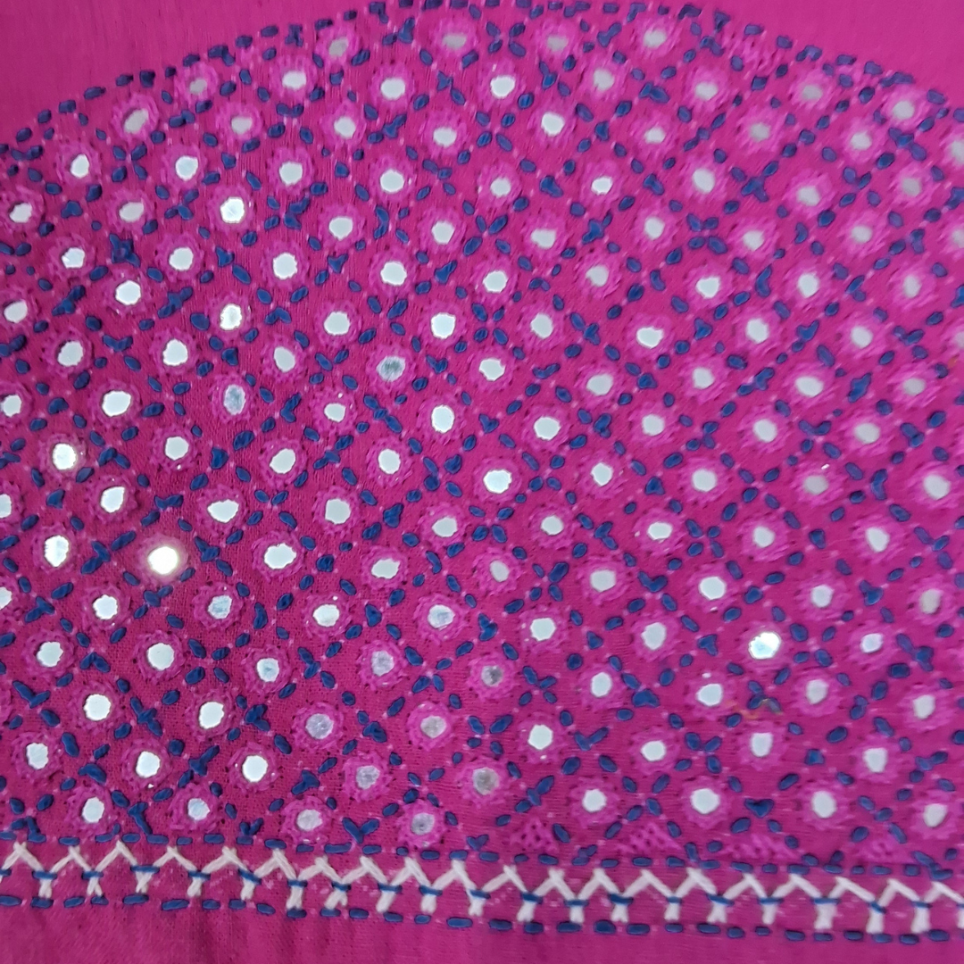 A close-up of a bright pink fabric with an all-over pattern of small white circular cutouts and dark blue stitched dots, plus a decorative border along the bottom edge.