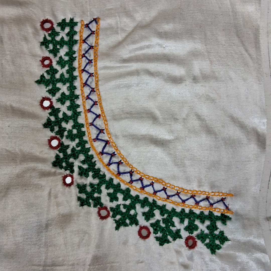A close-up of a cream-colored fabric with an embroidered curved neckline design featuring green floral embroidery, a yellow border, a purple lattice pattern, and small red and white mirror-work accents.