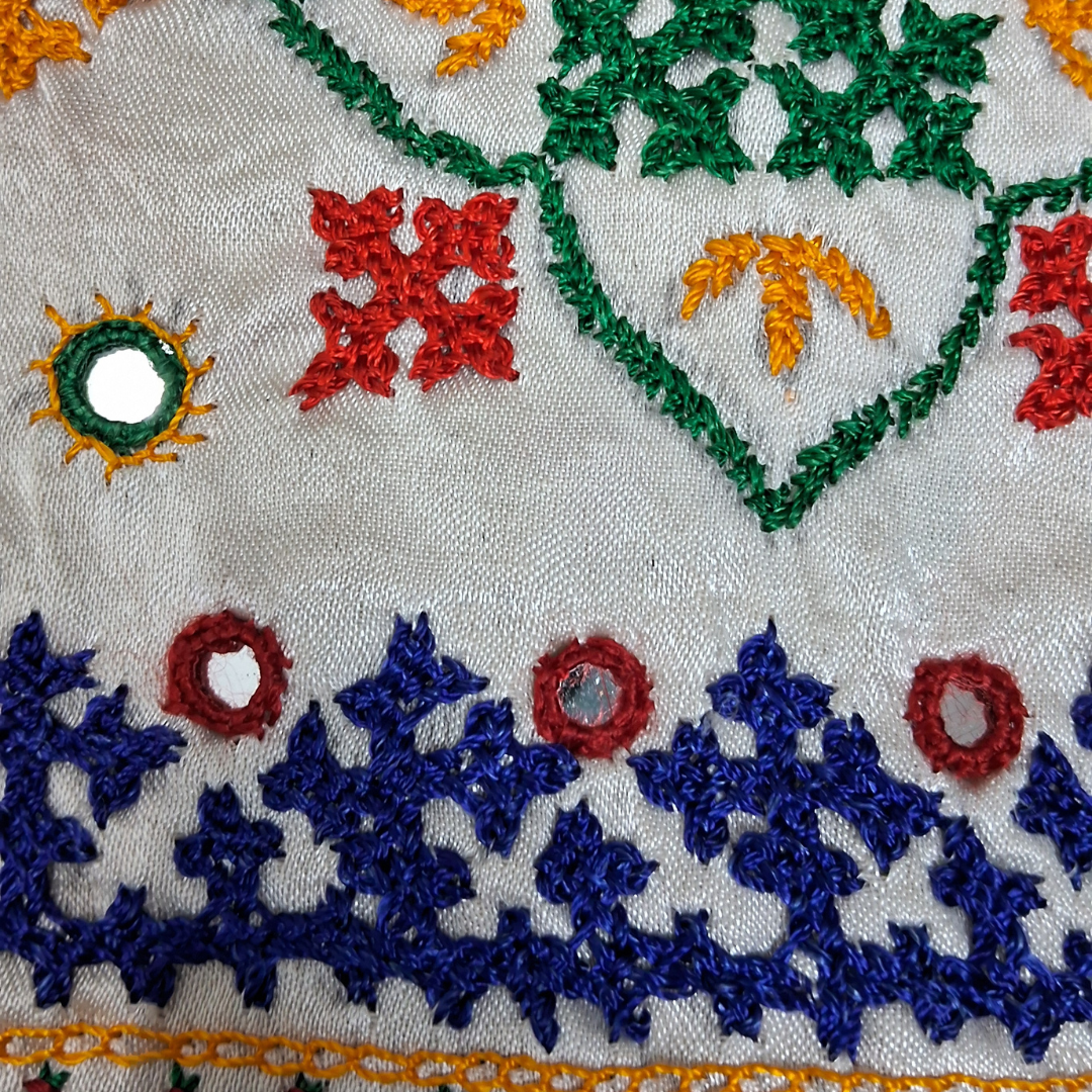 Close-up of an embroidered textile featuring multicolored floral and geometric stitching in green, blue, red, and yellow on a light fabric background, with small reflective mirror embellishments.